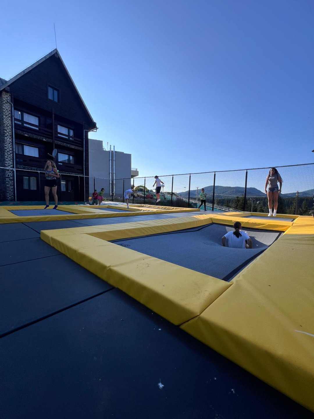 People bounce on interconnected yellow-bordered trampolines outdoors, with a wooden building on the left and mountains in the distance under a clear blue sky.
