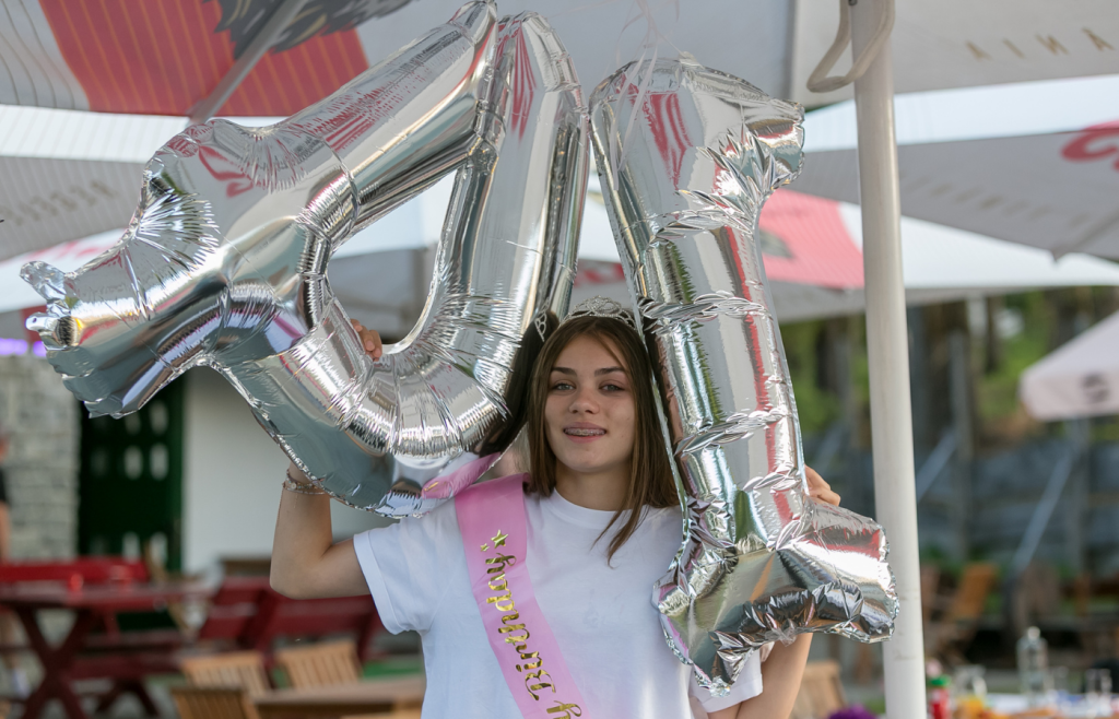 Young girl wearing a tiara and a pink sash at an outdoor birthday party, holding large silver foil number balloons overhead.