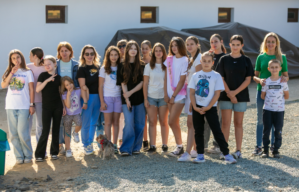 Group of children and adults posing for a photo outdoors, with a small dog at their feet in front of a building and covered objects in the background.