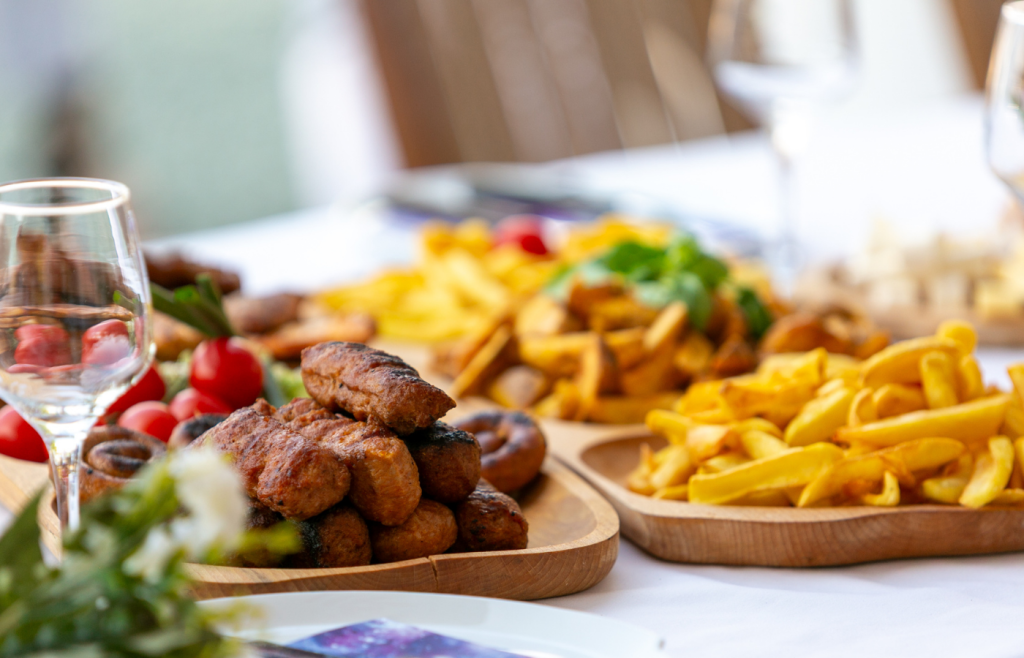 Stack of small grilled meatballs on a wooden platter with a pile of french fries, wine glass to the left.