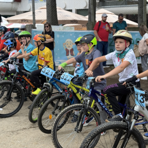 Kids on mountain bikes at a race start, wearing helmets with numbered bibs 321–326, ready to ride.