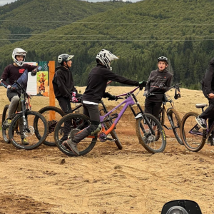 Group of young riders wearing helmets with mountain bikes on a sandy, open area and forested hills in the background.