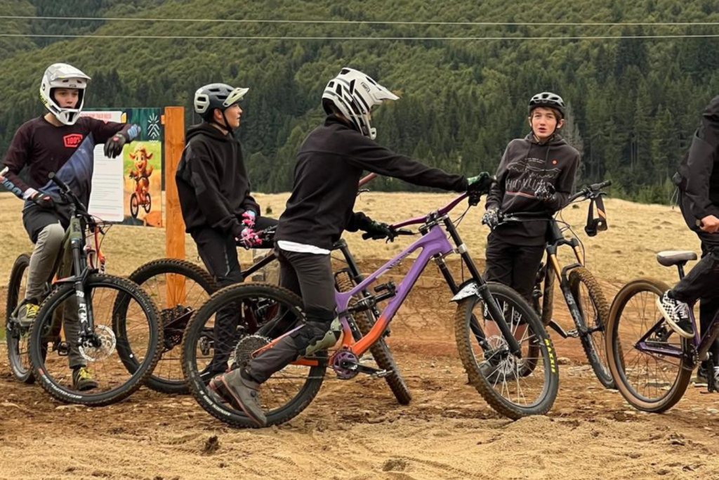 Group of young mountain bikers wearing helmets, standing with colorful bikes on a sandy trail with a forest backdrop.