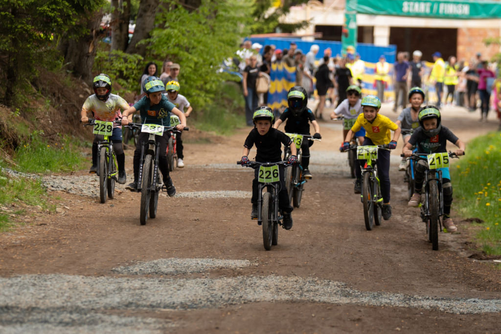 Young cyclists race along a dirt trail, helmets on, numbered bibs visible near the Start/Finish banner.