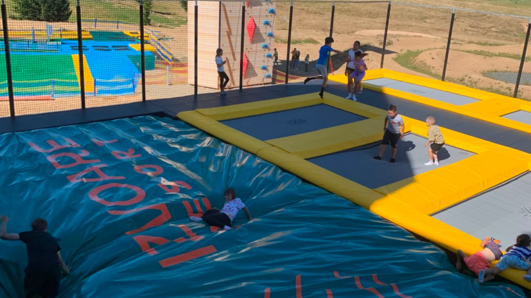 Children jump on a large indoor trampoline court with blue mats, yellow edges, and safety netting surrounding the area.